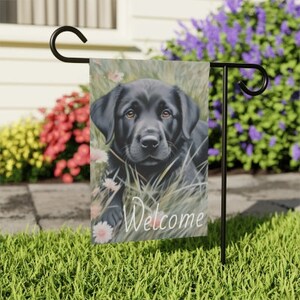 May include: A black Labrador Retriever puppy with brown eyes is lying in a field of green grass and white flowers. The words "Welcome" are printed in white on the flag.