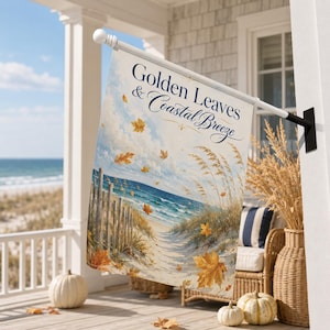 Puede incluir: Una bandera de jardín rectangular con una escena costera que muestra un camino de arena, una valla de madera y olas. El texto Golden Leaves & Coastal Breeze está impreso en azul oscuro. Hojas otoñales doradas cubren el cielo y la arena.