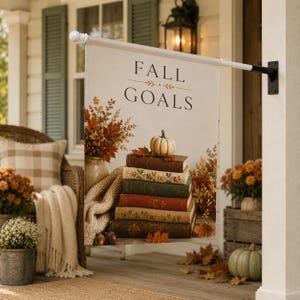 May include: A rectangular garden flag hanging from a black metal pole, featuring the text FALL GOALS in dark brown letters above a stack of five vintage books topped with a small white pumpkin, surrounded by autumn leaves and a beige knit blanket.