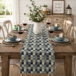 May include: A rectangular table runner featuring a blue, white, and cream patchwork pattern with floral and checkered squares. The runner is centered on a wooden dining table set with white ceramic bowls, wine glasses, and a white pitcher.