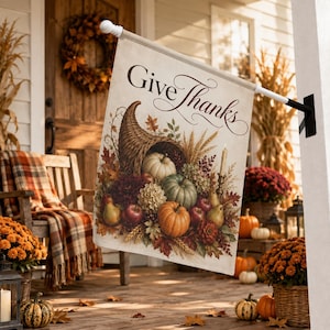 Könnte beinhalten: Eine rechteckige Gartenflagge mit dem Schriftzug Give Thanks in eleganter Schreibschrift. Das Motiv zeigt ein Füllhorn, das mit Kürbissen, Äpfeln, Birnen und herbstlichen Blättern in warmen Farben gefüllt ist, befestigt an einer Metallstange.