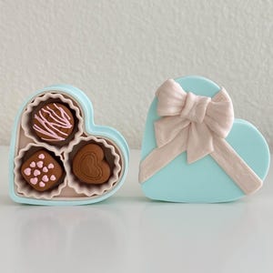 May include: A heart-shaped box of chocolates in light blue, with a matching lid featuring a pink bow. The open box reveals three chocolates with pink and white decorative icing.