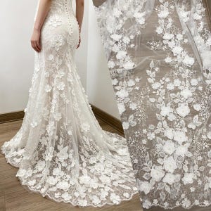 Puede incluir: Vestido de novia blanco con adornos florales. El vestido presenta una silueta de sirena con una larga cola y delicados apliques florales en 3D. La tela es un material transparente y de encaje, y el vestido tiene una espalda abotonada.
