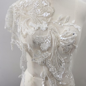 May include: White floral lace applique with sequins and beads. The applique features a delicate design of leaves and flowers, with intricate detailing. The lace is attached to a sheer fabric, creating a layered effect. Perfect for bridal wear or embellishments.