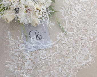 May include: Close-up of white lace fabric with floral and leaf patterns. The lace has a scalloped edge and is embellished with small sequins. A bouquet of white flowers is partially visible in the background.