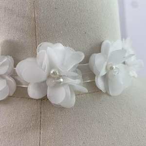 Może przedstawiać: Biały kwiatowy naszyjnik typu choker z delikatnymi kwiatami z tkaniny i perłowymi akcentami. Kwiaty są przymocowane do przezroczystej, elastycznej opaski, tworząc romantyczny i elegancki dodatek.