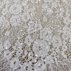 Puede incluir: Primer plano de tela de encaje blanco con patrones florales y lentejuelas dispersas. El delicado encaje presenta diseños intrincados y un borde festoneado, creando una apariencia texturizada y elegante. Ideal para ropa de novia o de ocasión especial.