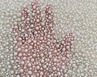 May include: Close-up of white beaded fabric with a hand underneath. The fabric is densely covered in small, round, white beads and sequins. The hand is visible through the sheer fabric, creating a layered effect. The beads vary in size, adding texture.