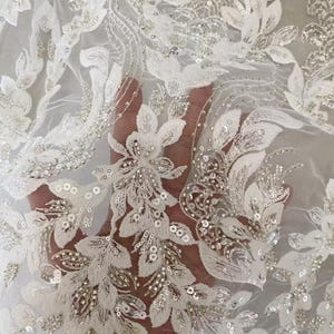 May include: Close-up of white lace fabric with floral and leaf patterns. The fabric is embellished with sequins and beads, creating a shimmering effect. The design features detailed embroidery and a delicate, sheer texture, suitable for bridal or formal wear.