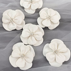 Può includere: Sei abbellimenti floreali in tessuto color avorio con un design a petali stratificati. Ogni fiore presenta un centro di strass scintillanti. I fiori sono disposti su uno sfondo di tessuto bianco trasparente.