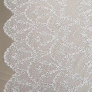 May include: Close-up of white lace fabric with a scalloped edge. The delicate lace features a floral pattern with intricate details. The fabric is likely used for bridal wear, dresses, or other elegant apparel.
