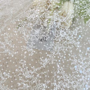 May include: Close-up of white lace fabric with floral patterns and scattered sequins. The delicate lace features intricate designs and a sheer appearance, ideal for bridal or formal wear. The sequins add a subtle sparkle to the fabric.