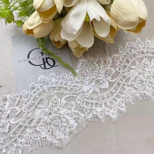 May include: White lace trim with pearl and sequin embellishments. The lace features a floral design and scalloped edges. A bouquet of white and yellow tulips is in the background, along with a card that says "WEDDING ACCESSORIES".