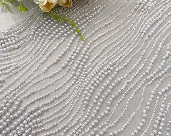 May include: Close-up of a white fabric adorned with rows of small, round, white pearls. The pearls are arranged in wavy lines across the fabric. A bouquet of cream-colored flowers and greenery is in the upper left corner.