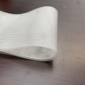 Peut inclure: Une section enroulée de tissu en maille blanche. La maille présente un motif de grille carrée fine. Le tissu est incurvé, montrant sa flexibilité. Le fond est une surface brun foncé.