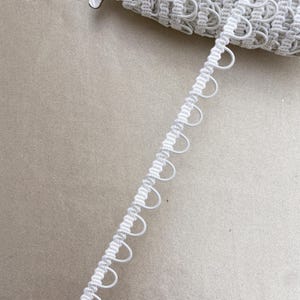 May include: White looped trim, ideal for sewing and crafting projects. The trim features a series of small, evenly spaced loops along one edge, creating a decorative border. The trim is rolled up, showing the texture and detail of the design.