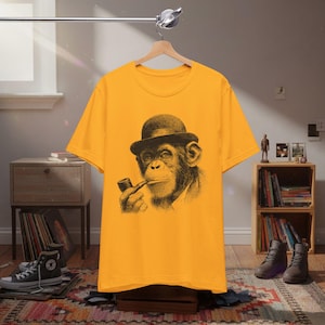 Puede incluir: Camiseta de color amarillo mostaza con una ilustración en blanco y negro de un chimpancé con bombín y pipa. La camiseta está colgada de una percha de madera.