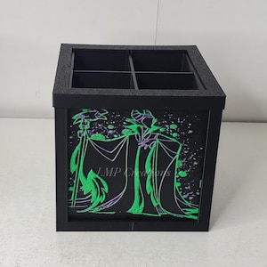 May include: A black square desk organizer with four compartments. The front panel features a green and purple graphic illustration of a character holding a staff, with the text LMP Creations printed in the center of the design.