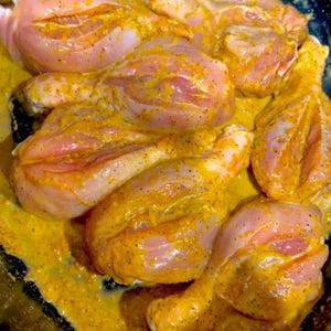 May include: Close-up of raw chicken legs marinated in a yellow sauce. The chicken is arranged in a black pan.
