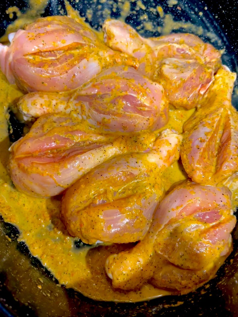 May include: Close-up of raw chicken legs marinated in a yellow sauce. The chicken is arranged in a black pan.