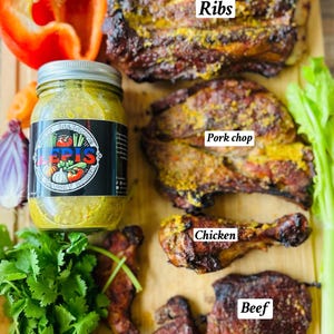 May include: A wooden cutting board with various types of meat, including ribs, pork chop, chicken, and beef. The meat is seasoned with a yellow spice and is ready to be served. There is a jar of green sauce and a red bell pepper on the board.