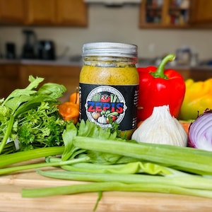 May include: A glass jar filled with a yellow sauce with a black label that says "ZEPIS" and has a picture of a garlic clove, a red pepper, a green pepper, and a tomato. The jar is sitting on a wooden cutting board with green celery stalks, green onions, a red onion, a white garlic clove, and a red bell pepper.