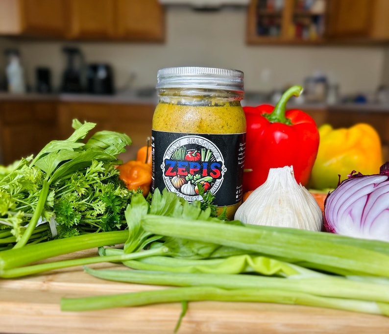 May include: A glass jar filled with a yellow sauce with a black label that says "ZEPIS" and has a picture of a garlic clove, a red pepper, a green pepper, and a tomato. The jar is sitting on a wooden cutting board with green celery stalks, green onions, a red onion, a white garlic clove, and a red bell pepper.