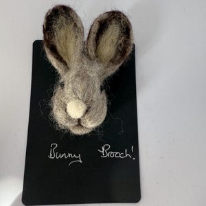 May include: A felt bunny brooch in shades of grey and brown, with long ears and a white nose. The brooch is mounted on a black card, with the words "Bunny Brooch!" written in white, perfect for adding a touch of whimsy.