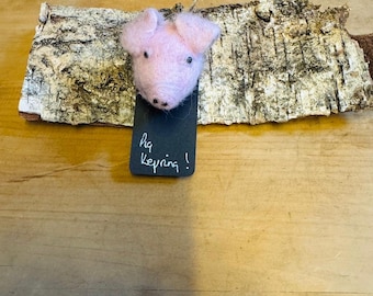May include: A pink felt pig head keychain hanging from a piece of birch bark with a black tag that says "Pig Keychain!"