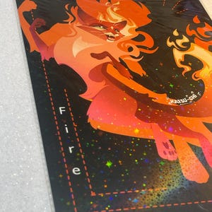 Pode incluir: Uma impress&atilde;o art&iacute;stica retangular apresentando um felino estilizado laranja e vermelho com pelo em forma de chamas contra um fundo escuro cheio de estrelas hologr&aacute;ficas. A palavra Fire est&aacute; impressa verticalmente em branco e o texto KATSU-CHI &eacute; vis&iacute;vel.
