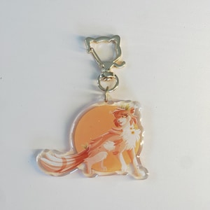 May include: A clear acrylic keychain featuring a stylized orange and white cat illustration set against a circular orange background. The cat has long fur, a bushy tail, and small white flowers on its chest. It includes a silver cat-shaped clasp.