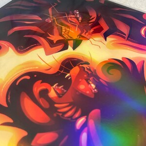 May include: A holographic art print featuring two stylized, dark-furred creatures with glowing orange and yellow accents. The top figure has sharp, angular features, while the bottom figure displays an open-mouthed expression with visible teeth.