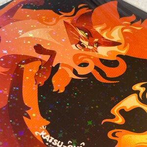 Pode incluir: Uma impress&atilde;o art&iacute;stica hologr&aacute;fica com um gato laranja estilizado com padr&otilde;es de pelo em forma de chama sobre um fundo escuro. O design inclui pequenas fa&iacute;scas em forma de estrela e o texto KATSU-CHI e _katsu_chi impresso em branco.