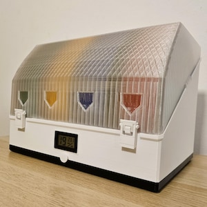 May include: A white, rectangular storage container with a clear, textured, angled lid. The container has four colored, triangular indicators and a digital display showing a percentage. The base is black.