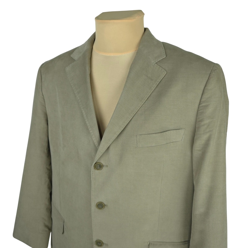 May include: A light green blazer with a three-button closure and a notched lapel. The blazer has a chest pocket and two lower pockets. The fabric has a subtle vertical texture. The blazer is displayed on a mannequin.