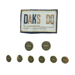 May include: A collection of gold-colored buttons and a label. The label is rectangular with the text "DAKS DO LONDON MADE IN GREAT BRITAIN" in navy blue and gold. The buttons have a repeating design.