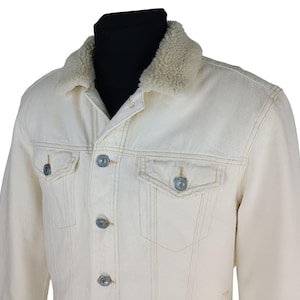 Puede incluir: Chaqueta vaquera color crema con cuello de borreguillo beige. La chaqueta tiene botones plateados y dos bolsillos en el pecho. Las costuras son de color beige en contraste.