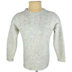 Puede incluir: Un suéter de manga larga en color gris claro con cuello redondo y un patrón de tejido texturizado. La zona superior del pecho presenta un detalle decorativo de punto calado que se extiende por los hombros y la parte superior de la espalda.