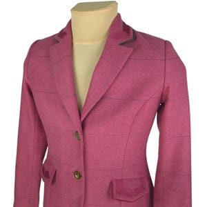 May include: A pink blazer with a herringbone pattern and a grid design. The blazer has a notched collar, two front pockets, and gold buttons. The blazer is displayed on a mannequin.