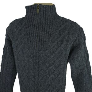 May include: A charcoal gray zip-up sweater with a high, ribbed collar. The sweater features a cable knit pattern across the body and sleeves. The zipper is a brass color. The sweater is made of a thick, warm material.