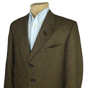 May include: A brown tweed blazer with a subtle plaid pattern in red and blue. The blazer has a notched lapel, two front pockets, and three black buttons. A light blue and white checkered dress shirt is visible underneath.