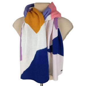 May include: A colorful scarf with a geometric pattern. The scarf features blocks of white, blue, orange, pink, and purple. The scarf is displayed on a mannequin. The scarf is made of a soft, knitted material.