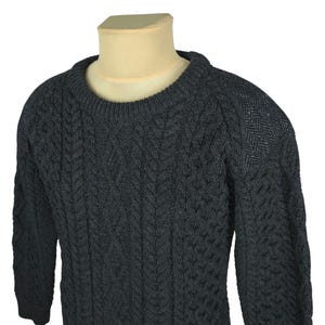 Puede incluir: Suéter de punto de cable gris oscuro con cuello redondo. El suéter presenta un diseño de punto de cable texturizado en la parte delantera y las mangas, con un patrón de espiga en los hombros. El suéter de manga larga se muestra en un maniquí.