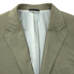 May include: Close-up of a tan corduroy blazer. The jacket has a notched lapel, a two-button closure, and a chest pocket. The interior lining is white. The fabric has a ribbed texture.