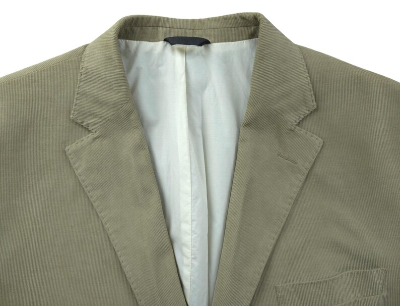 May include: Close-up of a tan corduroy blazer. The jacket has a notched lapel, a two-button closure, and a chest pocket. The interior lining is white. The fabric has a ribbed texture.