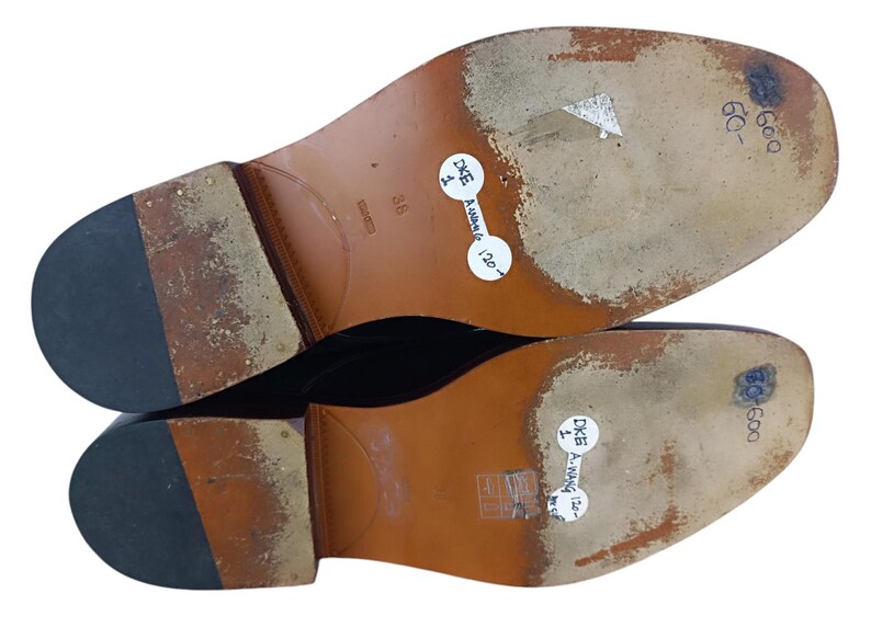 May include: Close-up of the soles of a pair of brown leather shoes. The soles are brown leather with a black rubber heel. The shoes are labelled "DKE" and "600".