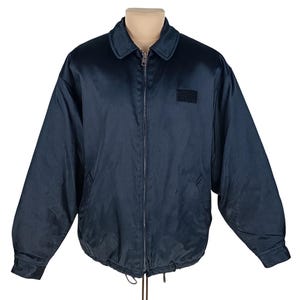 Op de afbeelding: Een donkerblauw jack met een ritssluiting en een klein patch op de linker borst. Het jack heeft een trekkoord aan de onderkant.