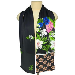 Puede incluir: Bufanda negra con diseño floral. La bufanda presenta una vibrante exhibición de flores rosas, blancas, azules y verdes sobre un fondo negro. La parte inferior de la bufanda tiene un patrón de pequeñas flores rosas.