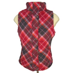 May include: A red, pink, and gray plaid vest with a high collar and button closure. The vest is sleeveless and quilted, with a subtle pink logo near the bottom hem. The pattern is a classic tartan design.