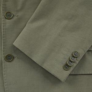 May include: Close-up of a sage green corduroy blazer. The blazer features three buttons on the sleeve cuff and two on the front. The fabric has a ribbed texture.